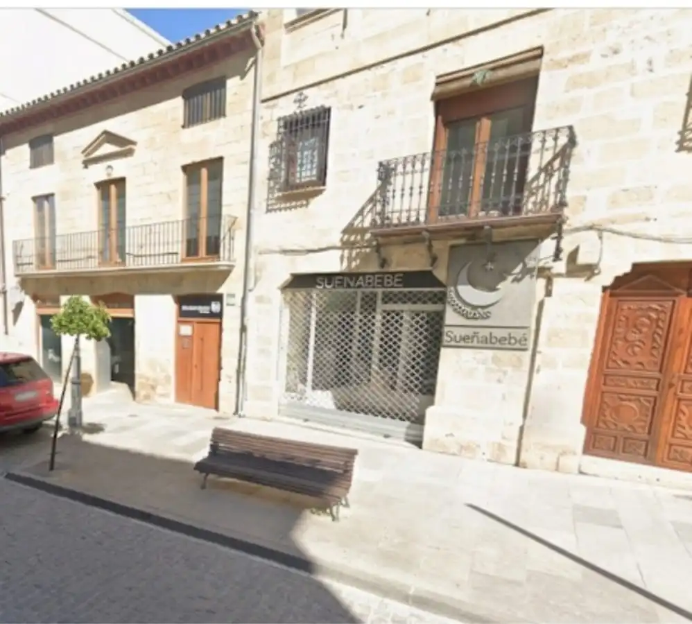 Esta foto captura una vista a nivel de calle de una calle histórica en Baeza, España, bajo la luz del día y un cielo azul despejado. Los edificios de la calle están hechos de piedra y madera tradicionales. El edificio principal presenta tres plantas y varios balcones con barandillas de hierro forjado. En la planta baja, hay una tienda de ropa para bebés y niños llamada "Sueño Bebe", con un cartel sobre la entrada. El local cuenta con un escaparate de cristal y una persiana metálica bajada. Hay un cartel con el logo de la tienda y el nombre "Sueño Bebe" a la derecha de la entrada de la tienda. Una banca de madera está ubicada frente a la tienda. Al lado del edificio principal, hay un edificio más pequeño de dos plantas con un balcón en el primer piso. Una puerta de madera marrón y un gran escaparate con la persiana metálica bajada se encuentran en la planta baja. Un automóvil rojo está estacionado en la calle.
