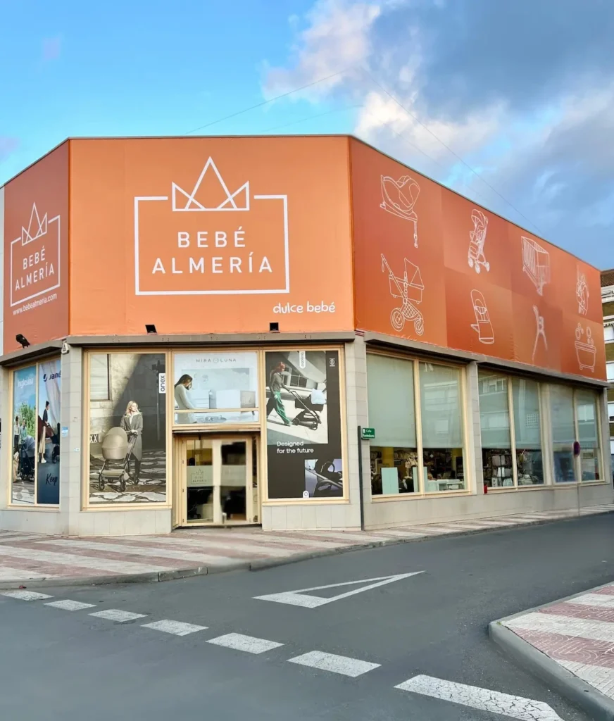 Fachada exterior de la tienda Dulce Bebé Roquetas de Mar, situada en una esquina con gran rótulo naranja. El cartel muestra el logo 'Bebé Almería' y pictogramas de carritos, tronas y cunas. Debajo, los grandes ventanales exponen productos de puericultura.